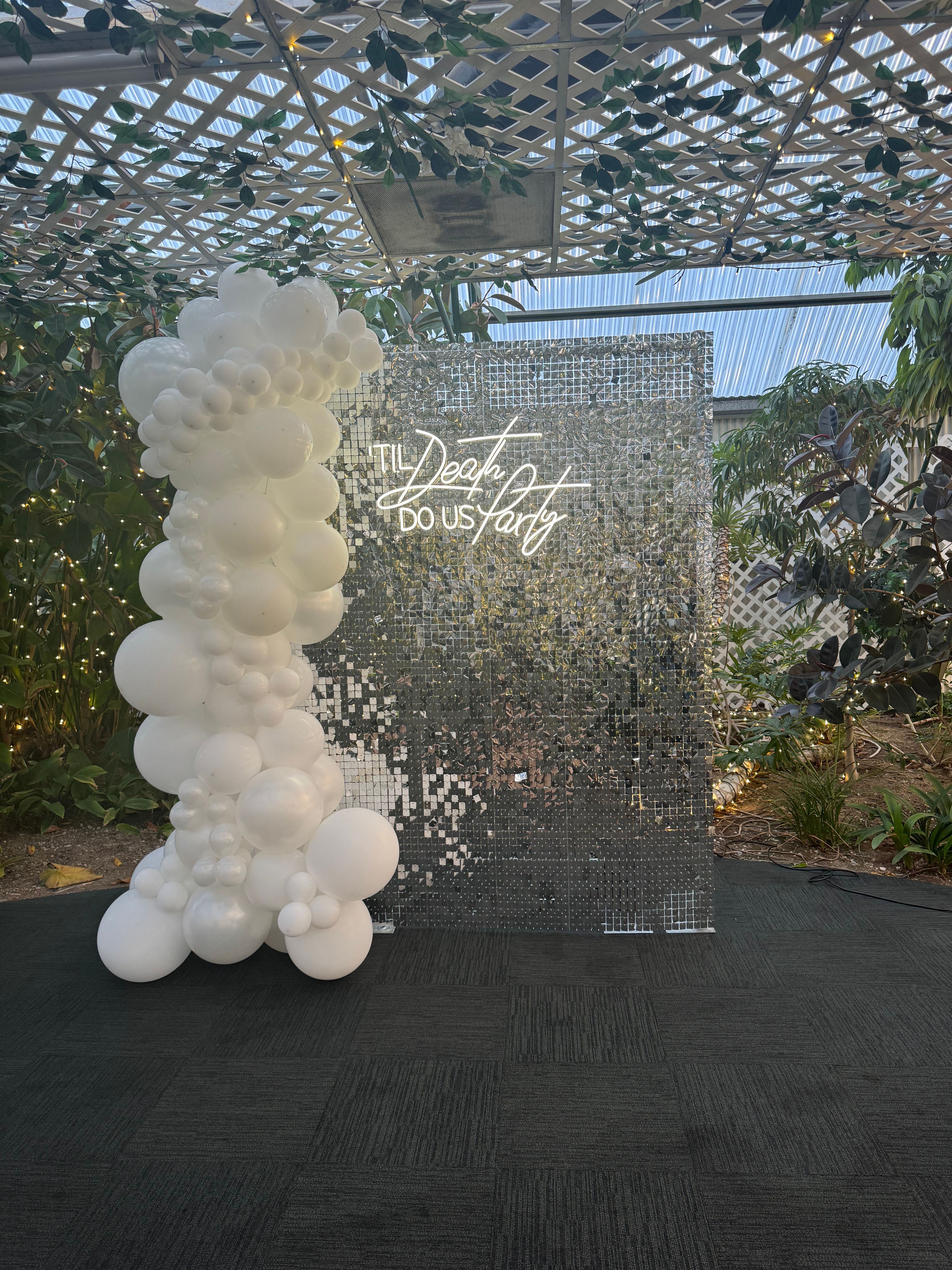 Playful wedding photo backdrop with a tall white balloon garland beside a mirrored sequin wall and neon sign reading "Til Death Do Us Party" under a leafy lattice canopy with string lights