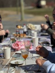 Sunny outdoor brunch table in a backyard setting with pastries on a tiered stand, crystal wine glasses, pink peony centerpiece and guests' hands reaching across the casual tablescape.