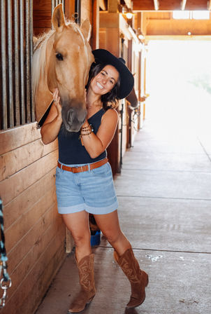 Smiling woman in a black hat and denim shorts hugging a palomino horse inside a sunlit wooden stable, wearing brown cowboy boots