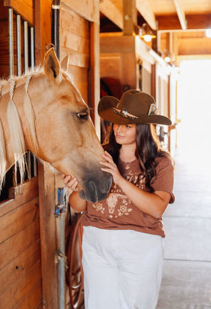 Cowgirl in a brown western hat gently stroking a braided-maned palomino horse inside a sunlit wooden stable aisle