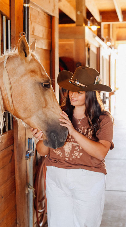 Cowgirl in a brown western hat gently stroking a braided-maned palomino horse inside a sunlit wooden stable aisle
