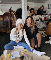 Two smiling women at a cozy indoor winter gathering, seated on a fur rug—one in a white fur hat and knit socks, the other in a black top; drinks, papers and a saddle visible in the rustic living room background.