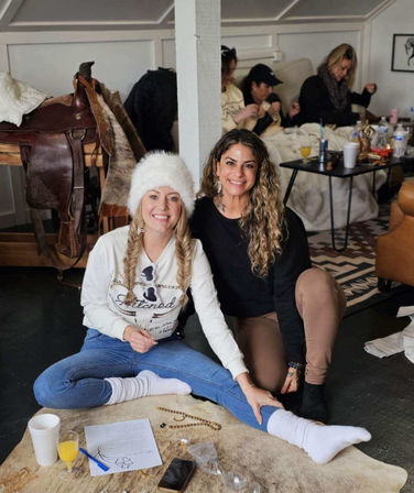 Two smiling women at a cozy indoor winter gathering, seated on a fur rug—one in a white fur hat and knit socks, the other in a black top; drinks, papers and a saddle visible in the rustic living room background.