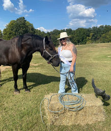 Person in a sun hat and sunglasses holding a dark horse by a lead rope in a sunny rural pasture, standing beside a hay bale with coiled lariat rope under a blue sky.
