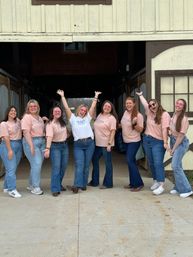 Eight friends in pink and white tees and jeans smiling and cheering at a rustic farm barn entrance.