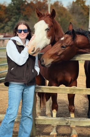 Person in sunglasses and denim leaning on a wooden fence petting two chestnut horses, one with a white blaze, in a sunlit paddock with autumn trees.