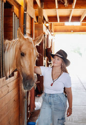 Woman in a cowboy hat and white tee petting a palomino horse in a sunlit wooden stable aisle at a ranch
