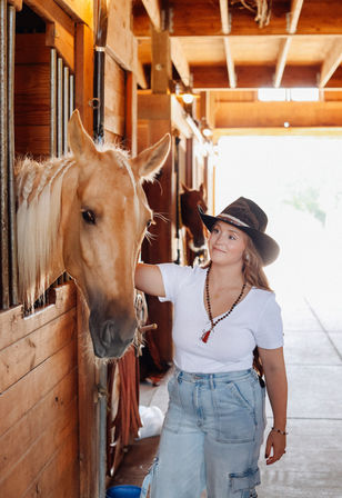Woman in a cowboy hat and white tee petting a palomino horse in a sunlit wooden stable aisle at a ranch