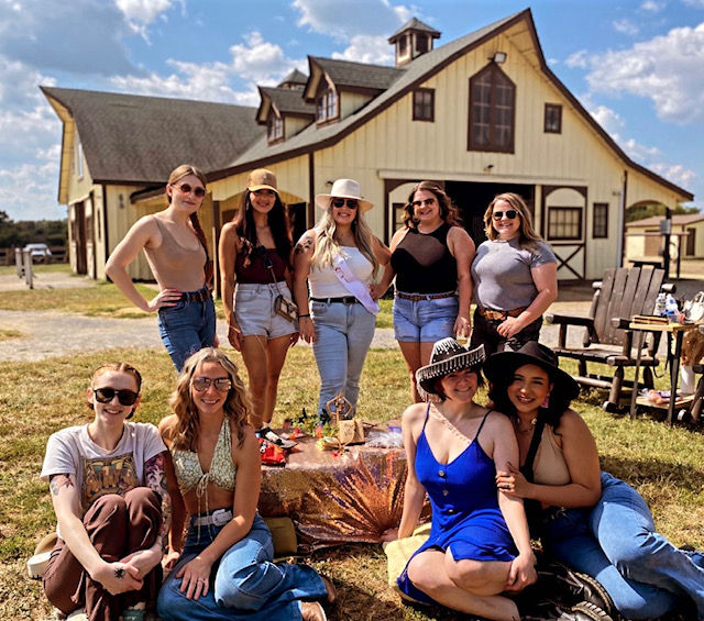 Nine women posing in front of a large yellow barn on a sunny day, seated and standing around a picnic setup on the grass, wearing casual summer outfits and hats for a cheerful country gathering.