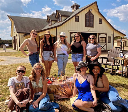 Nine women posing in front of a large yellow barn on a sunny day, seated and standing around a picnic setup on the grass, wearing casual summer outfits and hats for a cheerful country gathering.