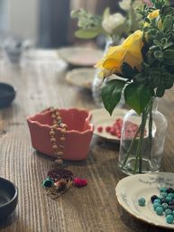 Farmhouse-style wooden table still life with a cheerful yellow rose in a glass vase, a pink ceramic bowl of wooden beads, and porcelain dishes holding turquoise beads and small red seeds.