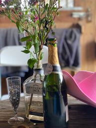 Flowers in a repurposed whiskey bottle on a rustic wooden table next to an unopened champagne bottle, a sequin-filled flute, and a bright pink cowboy hat in a cozy barn-like setting.