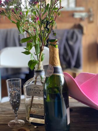 Flowers in a repurposed whiskey bottle on a rustic wooden table next to an unopened champagne bottle, a sequin-filled flute, and a bright pink cowboy hat in a cozy barn-like setting.