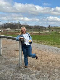 Smiling woman in denim jacket and 'BRIDE' T-shirt leaning on a metal rail at a country horse farm, holding a drink with green pastures, wooden paddocks, and a cloudy blue sky in the background.
