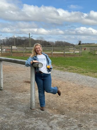 Smiling woman in denim jacket and 'BRIDE' T-shirt leaning on a metal rail at a country horse farm, holding a drink with green pastures, wooden paddocks, and a cloudy blue sky in the background.