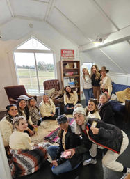 Group of women in cowgirl hats and cozy sweaters gathered in a bright barn-loft lounge with rugs, armchairs, a bookshelf and a large window overlooking a pasture; a "horses boarded" sign on the wall.