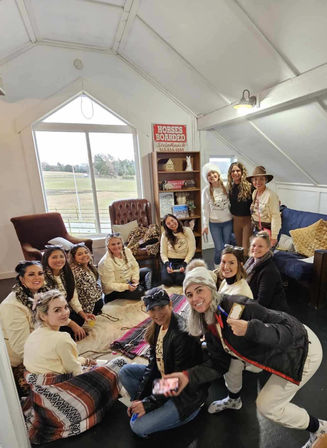 Group of women in cowgirl hats and cozy sweaters gathered in a bright barn-loft lounge with rugs, armchairs, a bookshelf and a large window overlooking a pasture; a "horses boarded" sign on the wall.