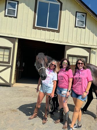 Three women in matching Nashville-themed tees, denim shorts and cowboy boots smiling with a dark horse outside a light-yellow barn on a sunny day