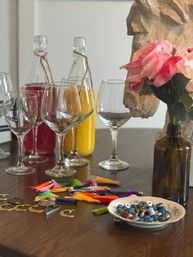 Wooden dining table styled with empty wine glasses, glass bottles of red and orange drinks, colorful tassel charms and metal pendants, a small dish of blue beads, and pink silk flowers in a brown bottle vase — casual boho tabletop decor.
