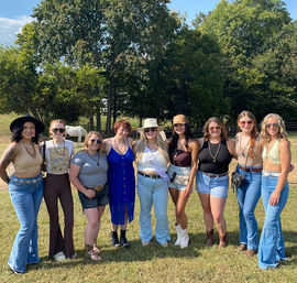 Nine women in country-chic outfits—jeans, boots, hats and sunglasses—posing in a sunny grassy pasture with trees and a white horse in the background.