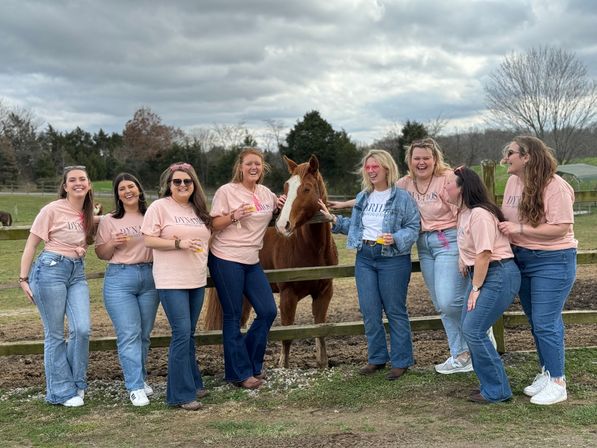 Eight women in matching pink shirts and jeans laughing and posing with a chestnut horse at a wooden fence on a country farm, green pasture and cloudy sky in the background.