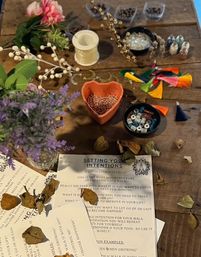 Overhead shot of a rustic wooden craft table set up for mala-making: heart-shaped bowl of pink beads, bowls of mixed beads, spool of string, colorful tassels, faux flowers and a 'Setting Your Intentions' worksheet scattered with dried leaves.