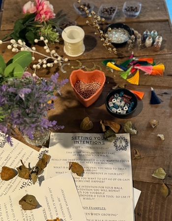 Overhead shot of a rustic wooden craft table set up for mala-making: heart-shaped bowl of pink beads, bowls of mixed beads, spool of string, colorful tassels, faux flowers and a 'Setting Your Intentions' worksheet scattered with dried leaves.