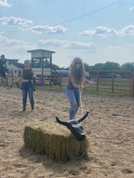 Two women in a sunny ranch arena practicing lassoing a black mock bull head mounted on a hay bale, with a horse, red corral fencing, barn structure and blue sky with clouds in the background.