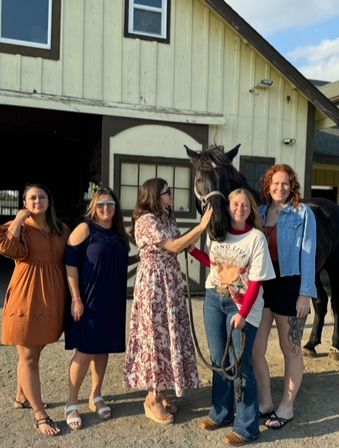 Group of five women smiling and posing with a black horse outside a cream-colored wooden barn — casual dresses, jeans and sandals in a sunny outdoor stable scene.