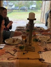 Rustic farmhouse table during a DIY beaded-rosary craft session with a brass candleholder, wine glasses, coffee mug, plates, and a blurred yard seen through an open barn door.