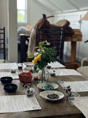 Sunlit loft craft table set for a DIY jewelry workshop: rustic wooden table with small bowls of gemstone beads, stringing supplies, printed instructions and a yellow rose centerpiece.
