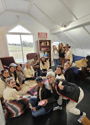 Smiling group of women gathered on blankets in a cozy ranch loft lounge with rustic bookshelf, a red "Horses Boarded" sign and a large window overlooking a pasture