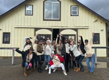 About a dozen women in matching cream shirts and winter jackets striking playful poses and cheering in front of a pale yellow barn on an overcast rural day, many wearing leopard print, boots and sunglasses with puddles on the gravel.