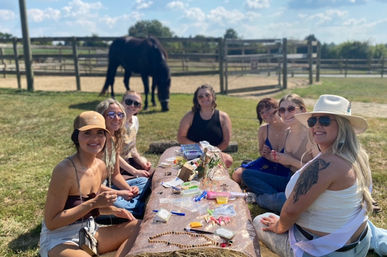 Group of women crafting on a hay-bale picnic at a sunny horse farm, seated around a makeshift table with beads and supplies, horse grazing behind a wooden fence