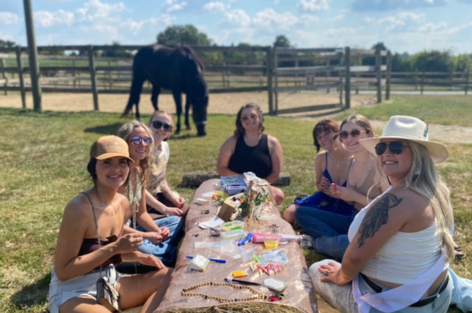 Group of women crafting on a hay-bale picnic at a sunny horse farm, seated around a makeshift table with beads and supplies, horse grazing behind a wooden fence