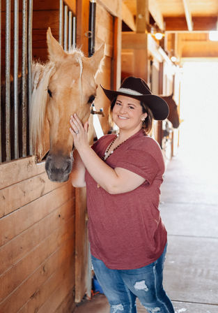 Smiling woman in a cowboy hat petting a palomino horse inside a sunlit wooden barn aisle — casual jeans and tee, country stable equestrian portrait.