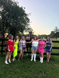 Six women in colorful dresses and boots pose with a brown horse at a wooden fence on a rural farm at dusk, grassy pasture with trees and a red barn in the background.