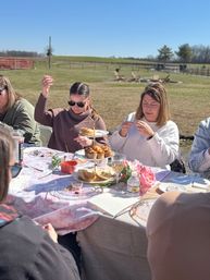 Sunny countryside outdoor tea party in a rural field with friends around a table set with a tiered stand of pastries, plates, flowers and drinks.