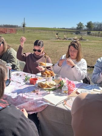 Sunny countryside outdoor tea party in a rural field with friends around a table set with a tiered stand of pastries, plates, flowers and drinks.