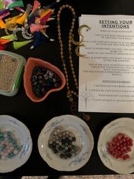 Overhead of a DIY mala bead workshop on a dark table: an intentions instruction sheet titled Setting Your Intentions, a wooden mala strand with gold crescent charms, colorful tassels, a heart bowl of patterned beads, and gemstone beads sorted in three floral saucers.