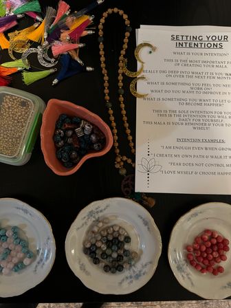 Overhead of a DIY mala bead workshop on a dark table: an intentions instruction sheet titled Setting Your Intentions, a wooden mala strand with gold crescent charms, colorful tassels, a heart bowl of patterned beads, and gemstone beads sorted in three floral saucers.