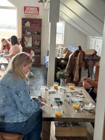 Cozy farmhouse loft craft session with a wooden table covered in beads, tools and glasses of orange juice, saddle decor and people crafting in the background