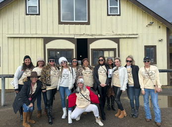 Cheerful group of women wearing matching beige shirts, winter jackets, hats and boots posing together in front of a rustic yellow wooden barn on an overcast day.