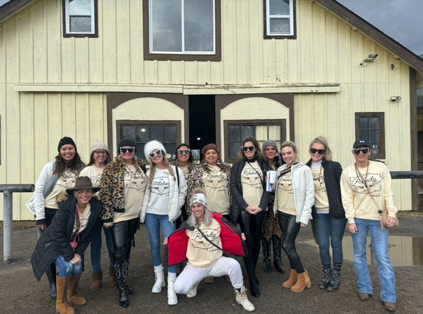 Cheerful group of women wearing matching beige shirts, winter jackets, hats and boots posing together in front of a rustic yellow wooden barn on an overcast day.