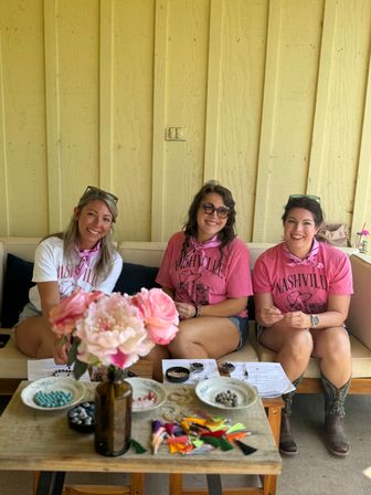 Three smiling friends in matching pink "Nashville" shirts and bandanas seated on a bench against a yellow wooden wall, with a table in the foreground holding a vase of pink flowers, beads and colorful tassels for crafting.