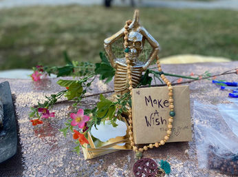 Outdoor bridal-wishing table with a playful gold skeleton figurine draped in wooden beads, handwritten "Make a Wish" card, pink flowers and a rose-gold sequin tablecloth.