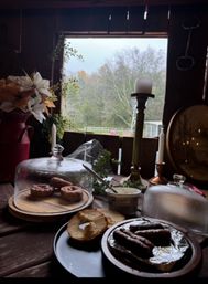 Cozy rustic cabin breakfast scene on a wooden table with glass-cloche-covered pastries, bagel halves, a plate of sausages, a tub of spread and candlesticks by an open window overlooking a misty autumn woodland.