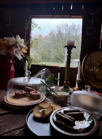 Cozy rustic cabin breakfast scene on a wooden table with glass-cloche-covered pastries, bagel halves, a plate of sausages, a tub of spread and candlesticks by an open window overlooking a misty autumn woodland.