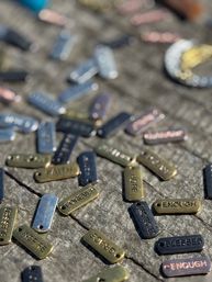 Sunlit close-up of scattered metal word charms on weathered wood — small rectangular tags stamped with inspirational words like HOPE, FOREVER, ENOUGH, and BLESSED in mixed brass and dark finishes.