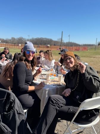 Smiling group of friends seated at a long outdoor table in a sunny grassy field, wearing beanies and sunglasses, enjoying drinks, snacks and a casual craft activity.